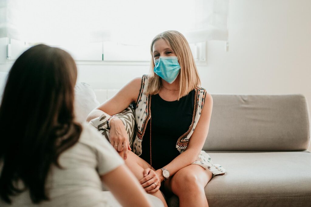 A woman wearing a medical mask sits on a couch talking to another woman.